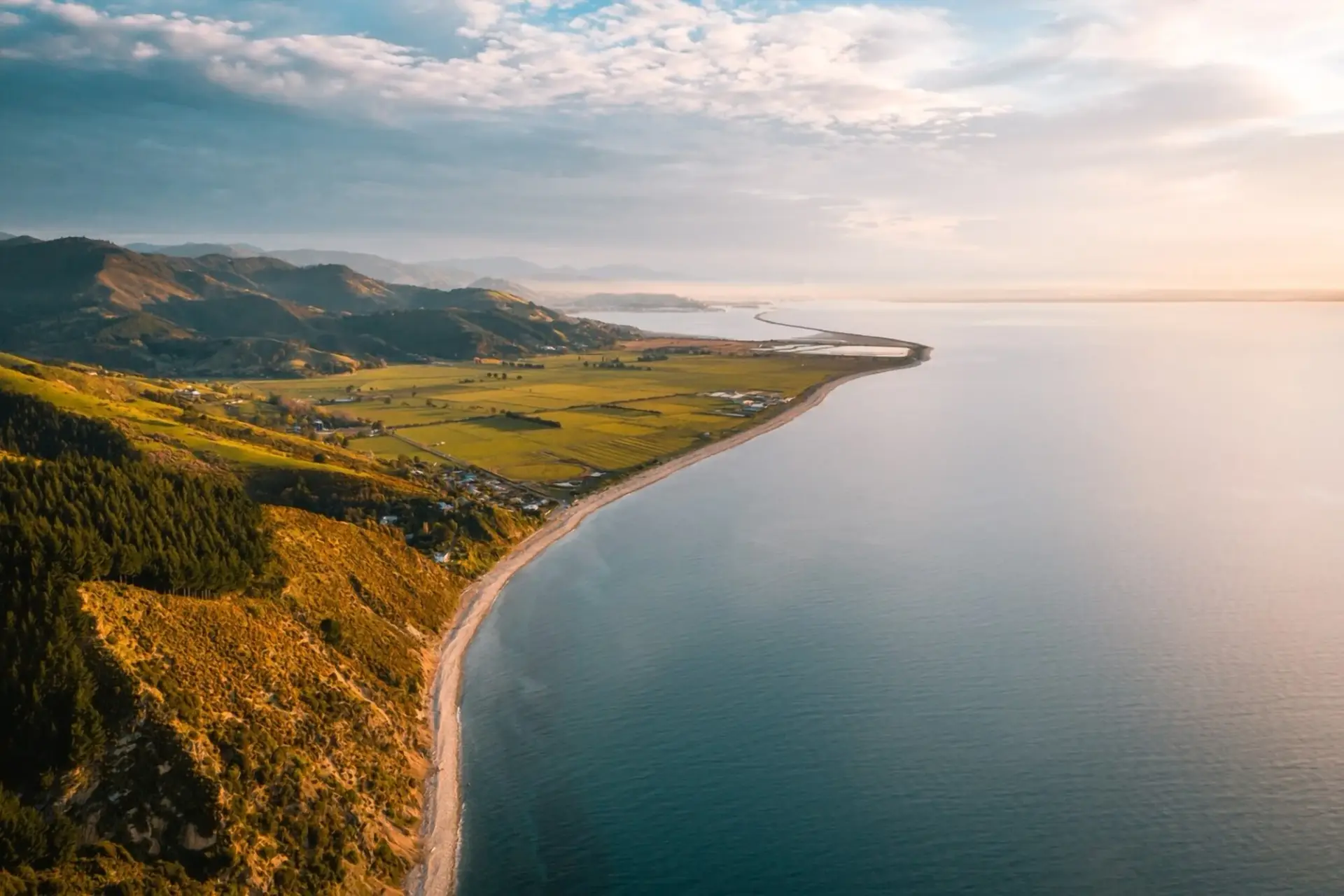 The Ruby Bay and Motueka coastline from the air at first light: a long shingle beach curving past orchards and paddocks, with bush hills on one side and Tasman Bay on the other.